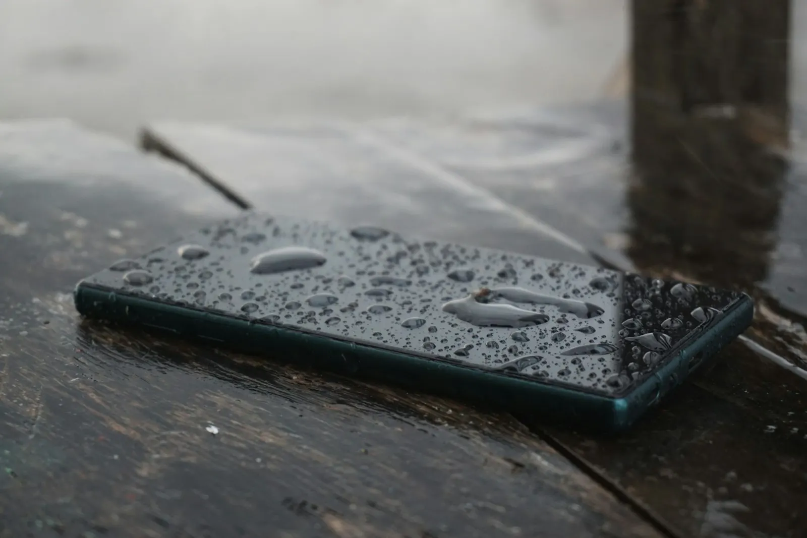 A smartphone covered in water droplets on a wet wooden surface