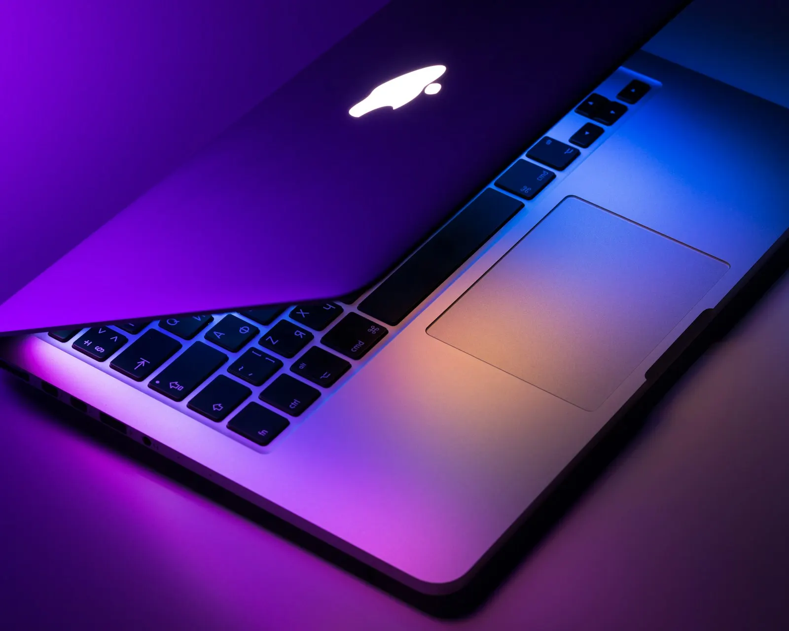 A MacBook opened slightly with its keyboard lit under colored light