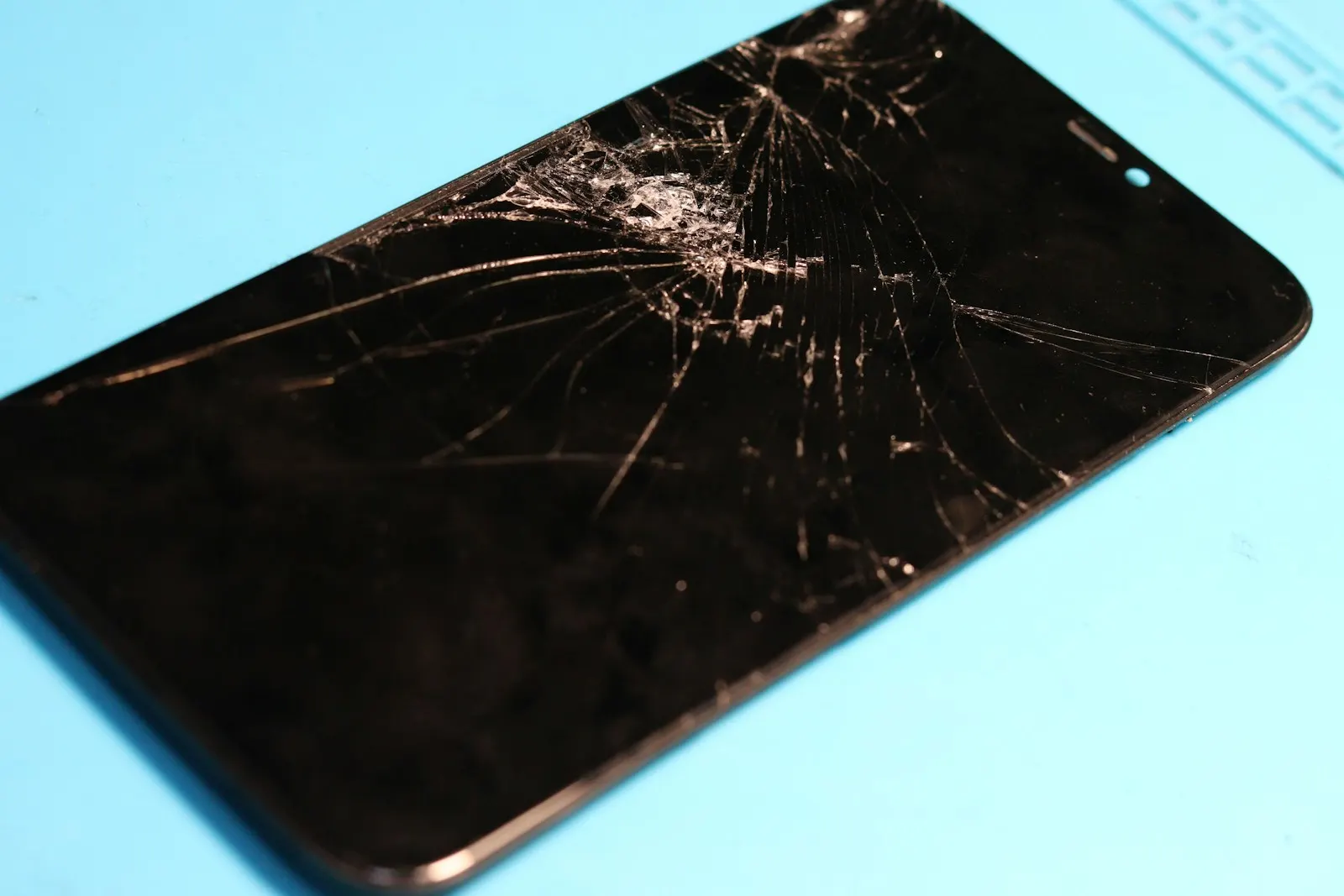 A phone with a shattered screen lying on a blue repair mat
