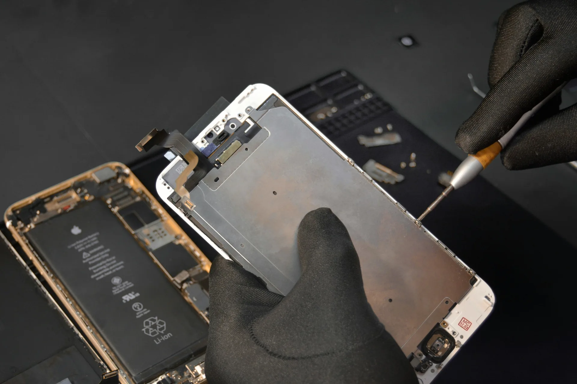 Gloved hands lifting the display assembly away from an opened iPhone on a dark bench, its battery and screws exposed
