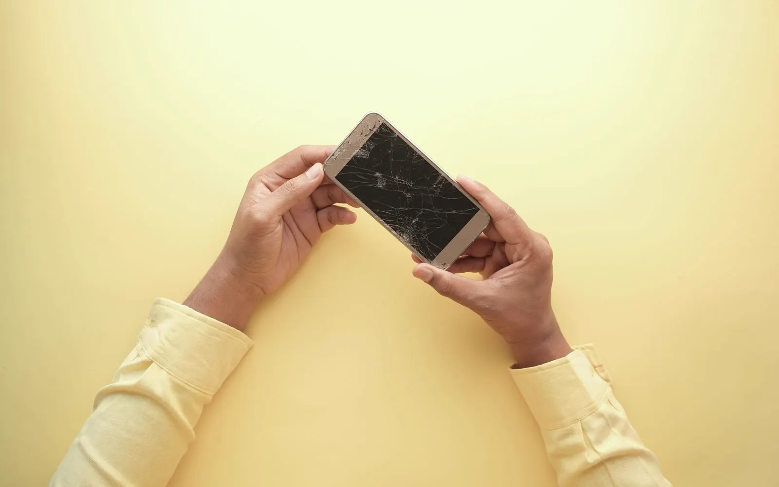 Two hands holding a cracked smartphone against a yellow backdrop