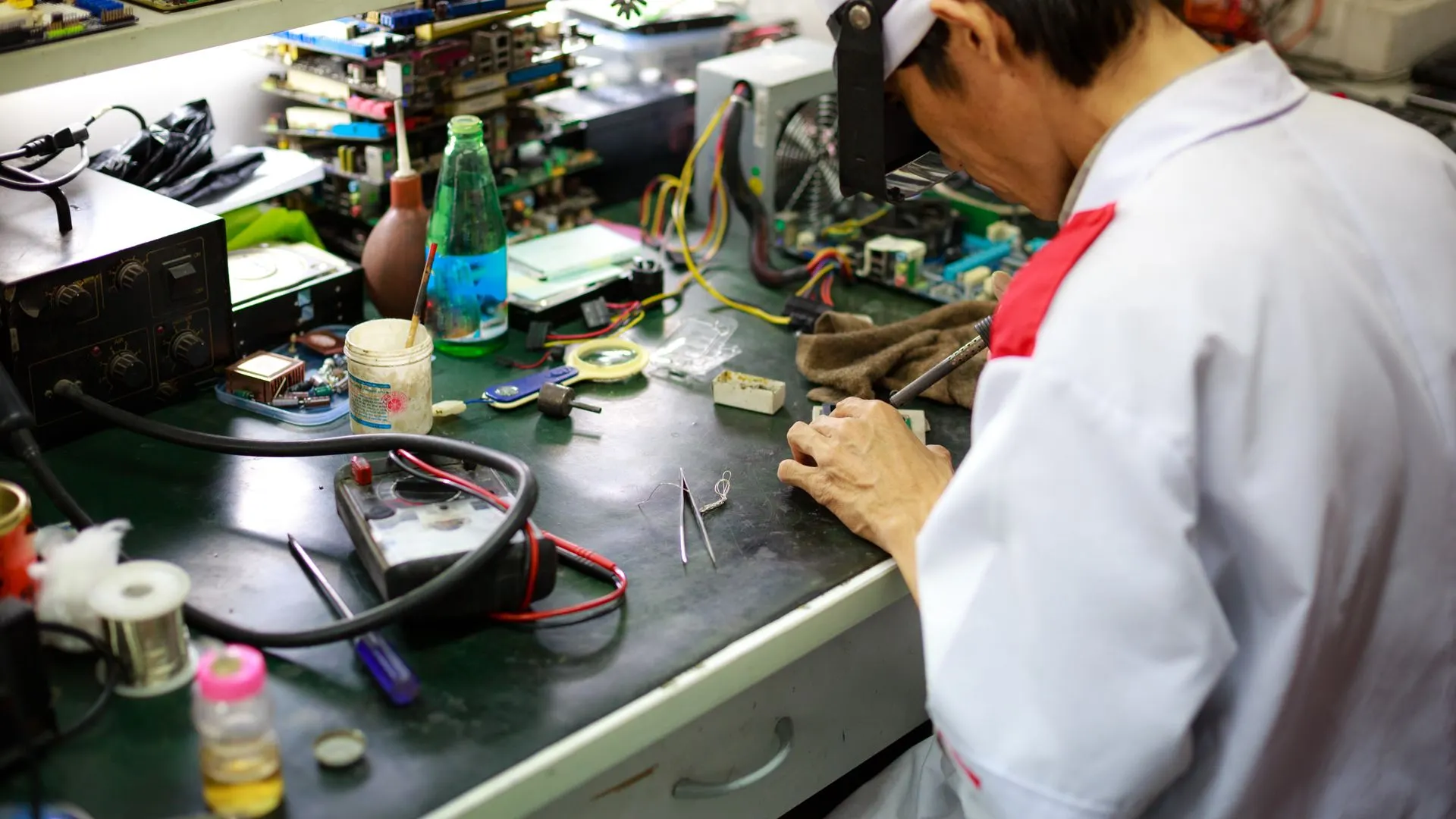 A repair technician in a white coat working at an electronics bench crowded with circuit boards, wires and soldering tools