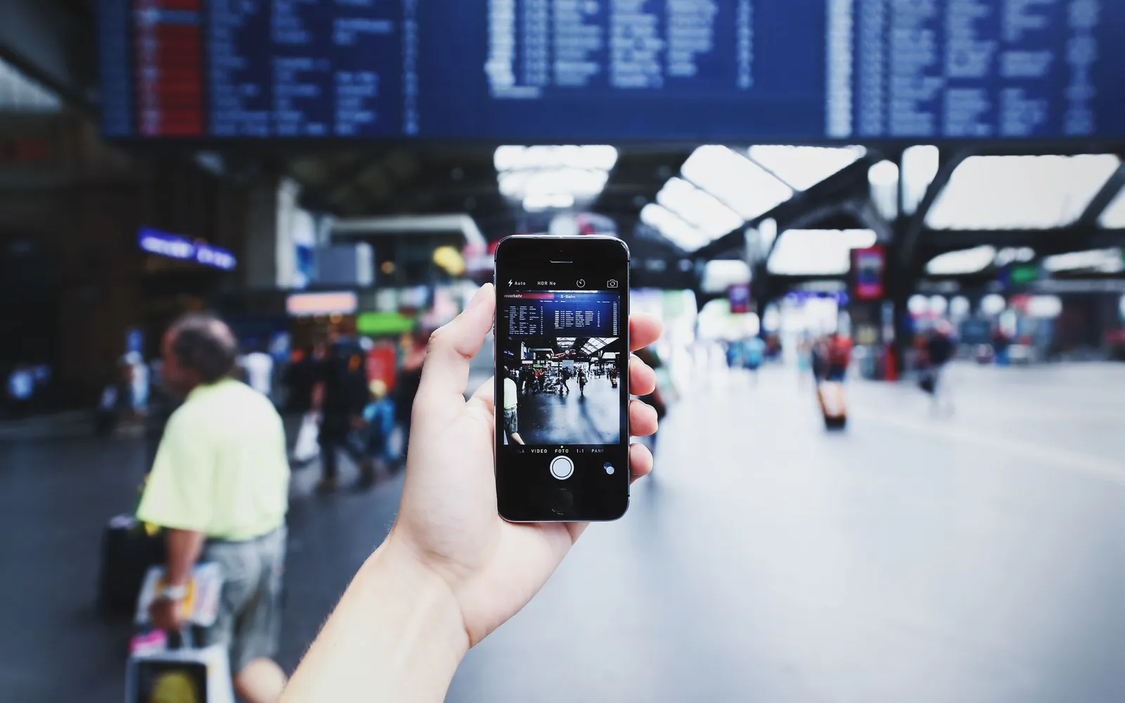 A hand holding up an iPhone to photograph a busy station hall under a departures board