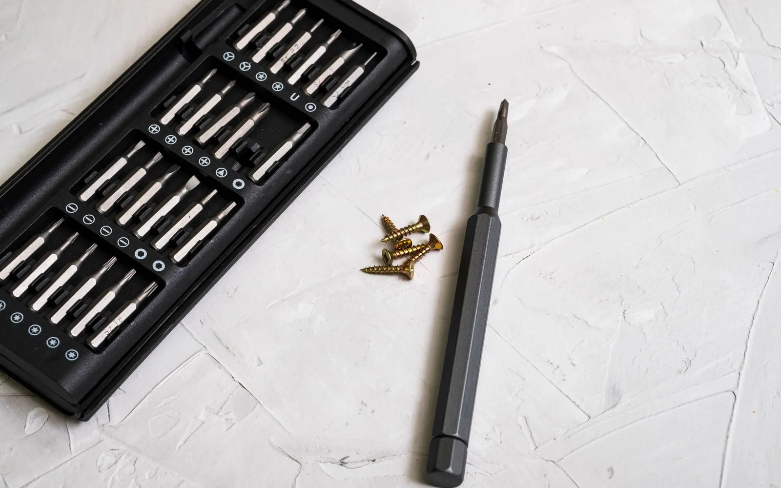 A precision screwdriver and its bit set beside a few small screws on a white bench