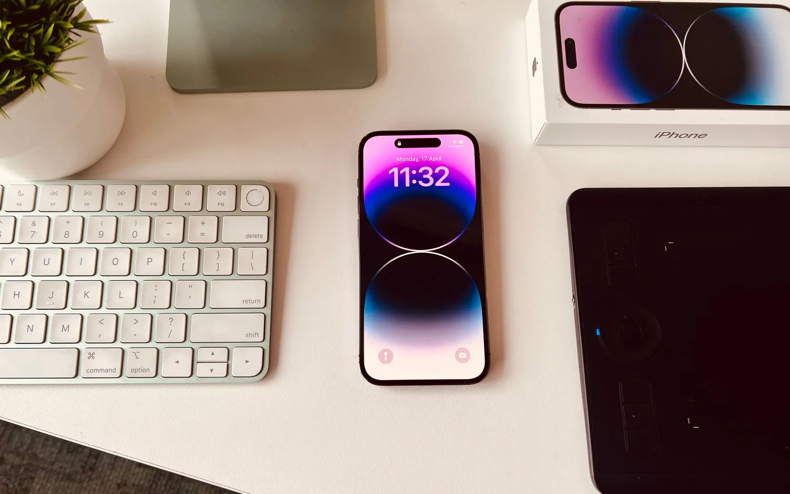 An iPhone Pro with its lock screen lit, on a white desk between a keyboard, a tablet and its box