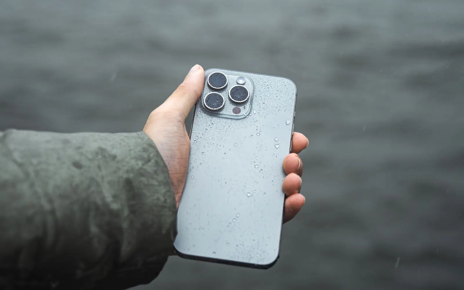 A hand holding an iPhone Pro, its back glass covered in raindrops