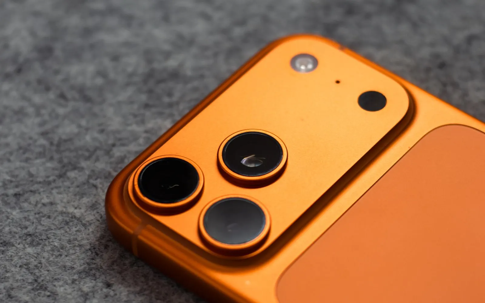 The triple rear camera of an orange iPhone Pro lying on grey felt