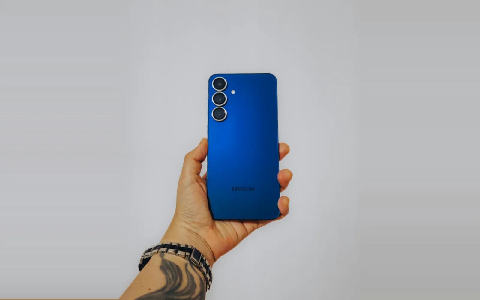 A hand holding up a blue Samsung Galaxy phone against a pale grey wall, its three rear cameras facing out