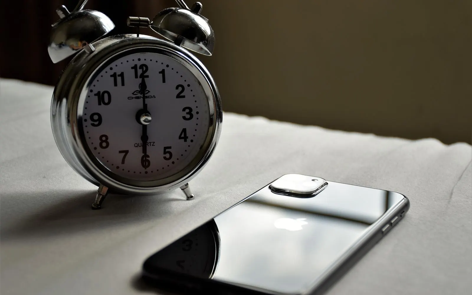 A silver alarm clock standing beside a smartphone lying face down on a table