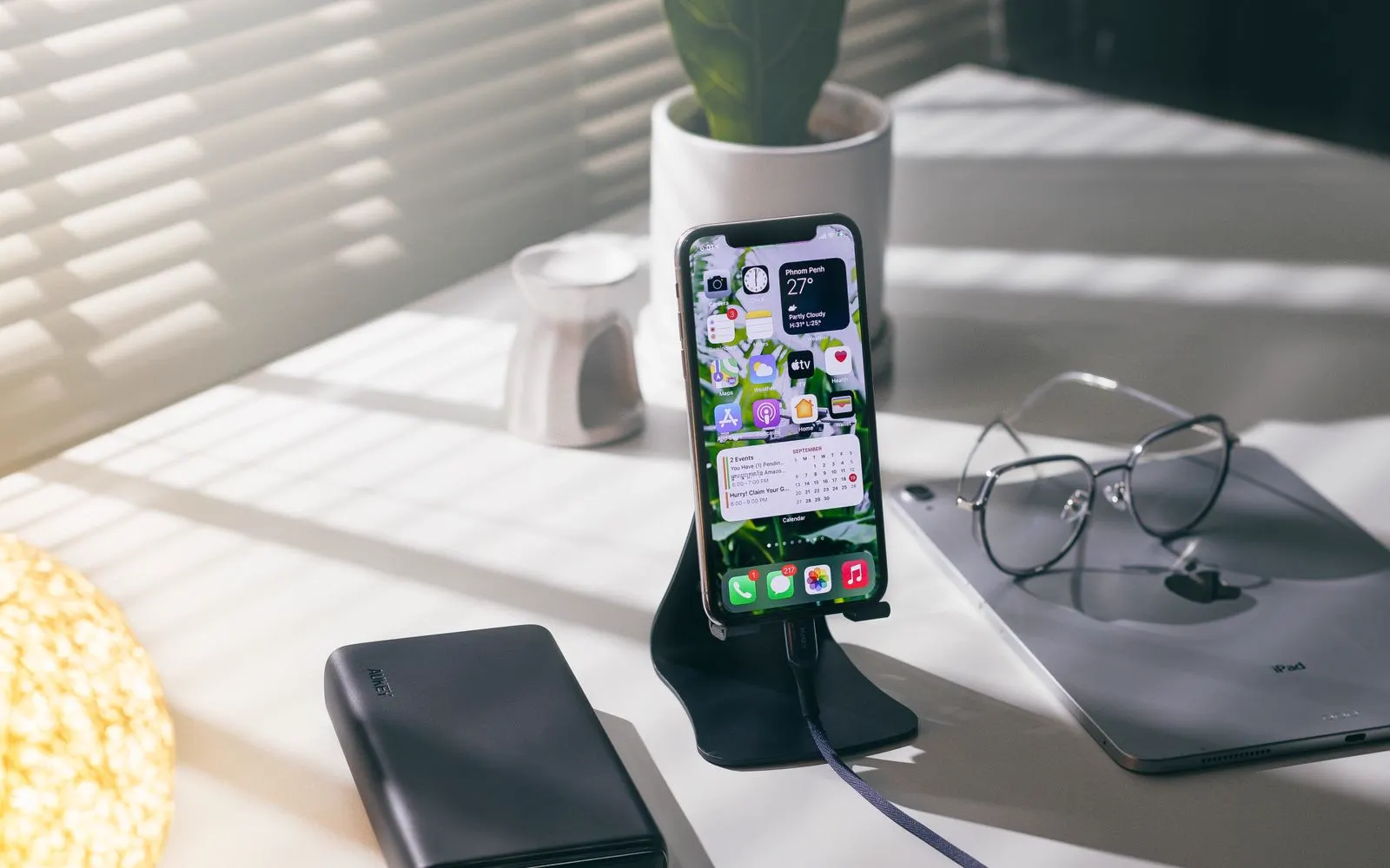 A device charging on a stand beside a power bank and an iPad on a sunlit desk
