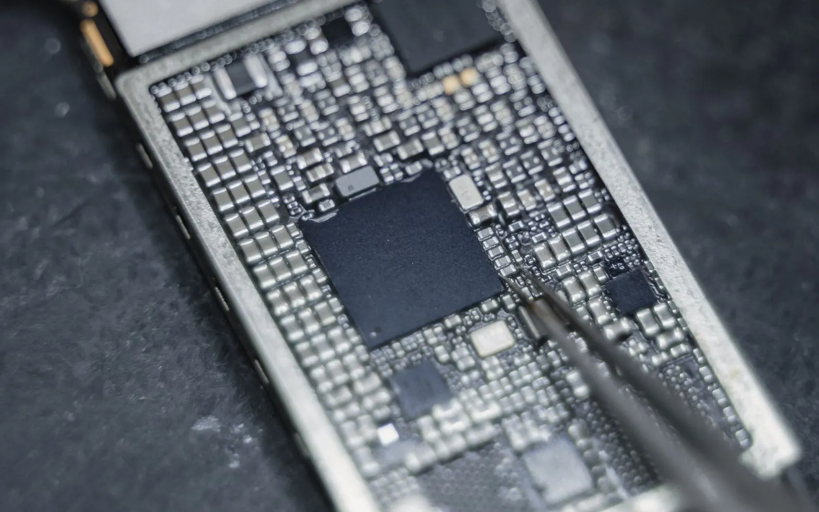 Tweezers working on a bare smartphone logic board on a dark mat