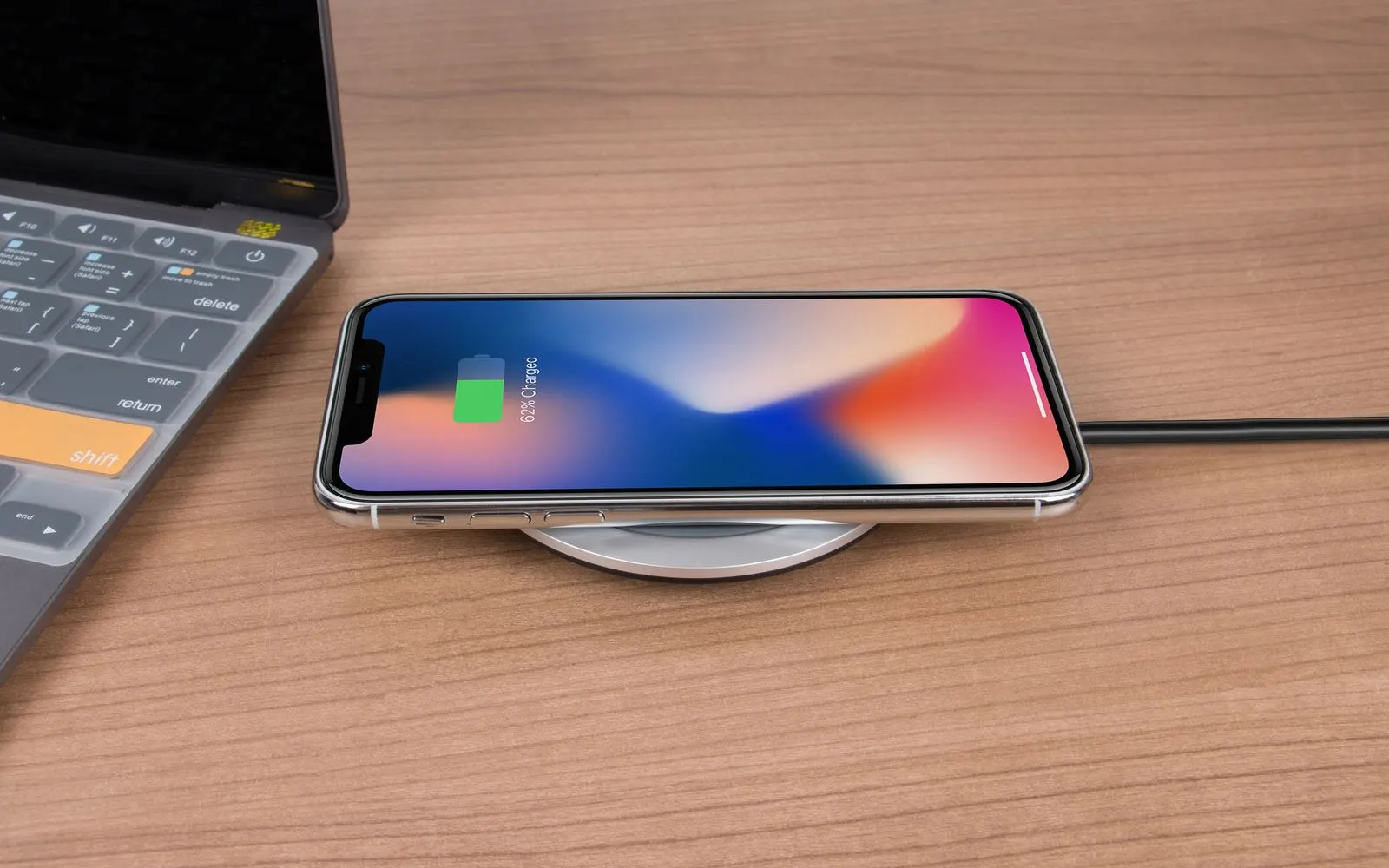An iPhone charging on a wireless pad beside a laptop, its screen showing the charge level