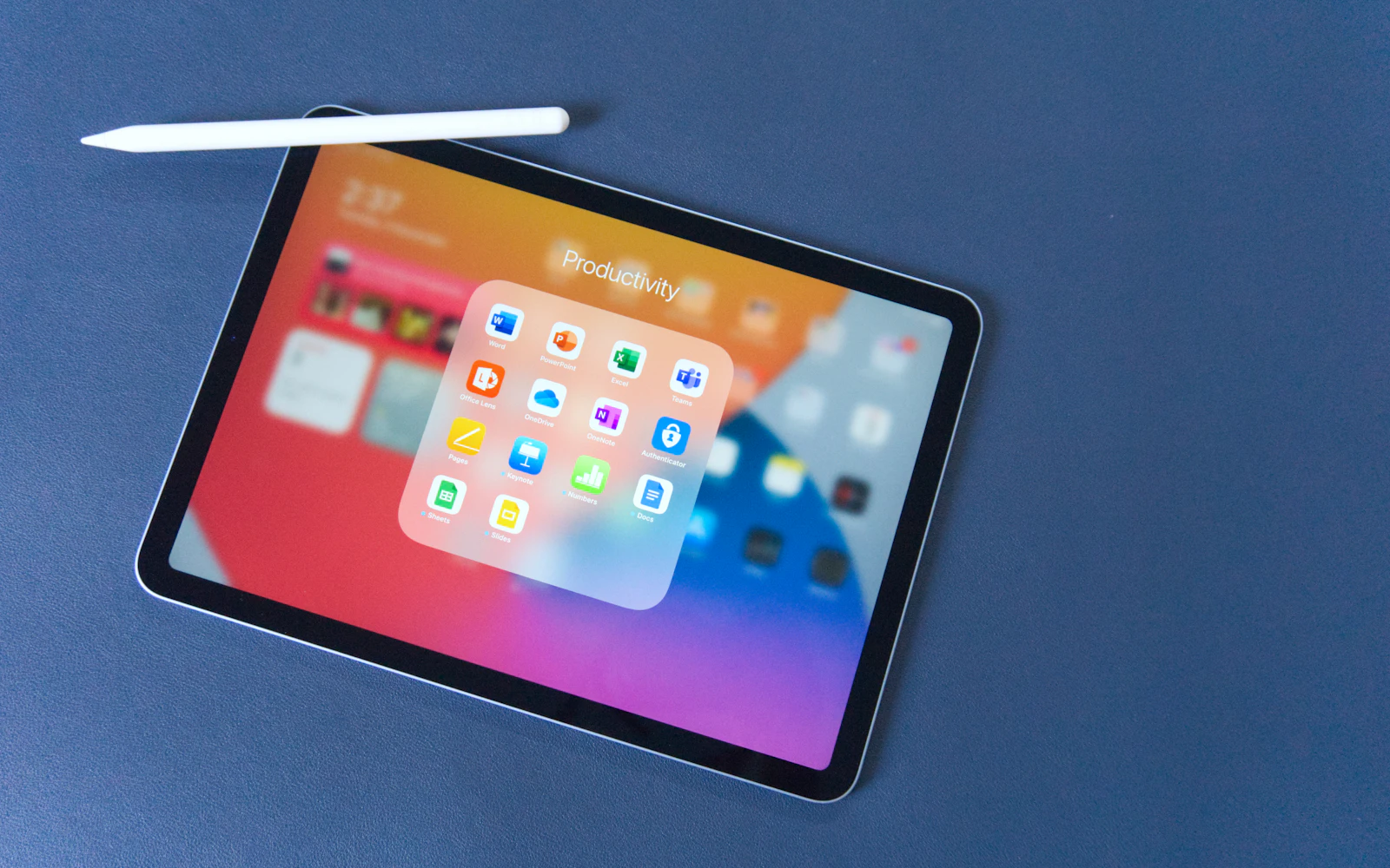 An iPad with an Apple Pencil on a blue surface, its home screen lit
