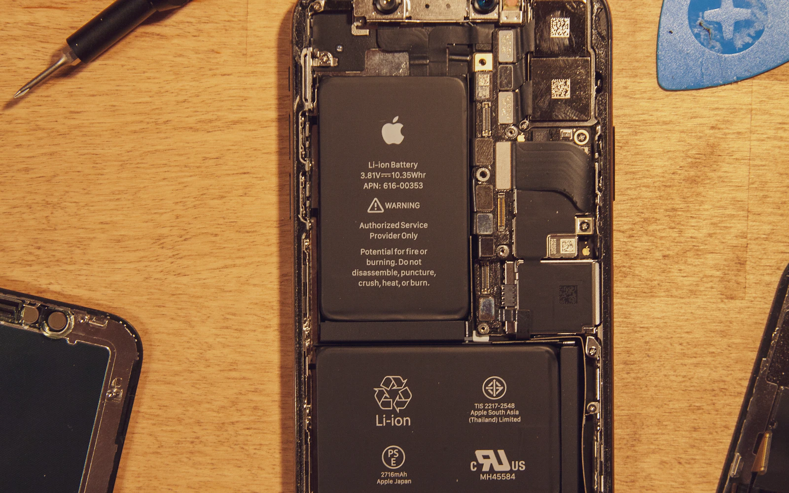 An opened iPhone on a wooden bench, its Li-ion battery and internals exposed beside repair tools