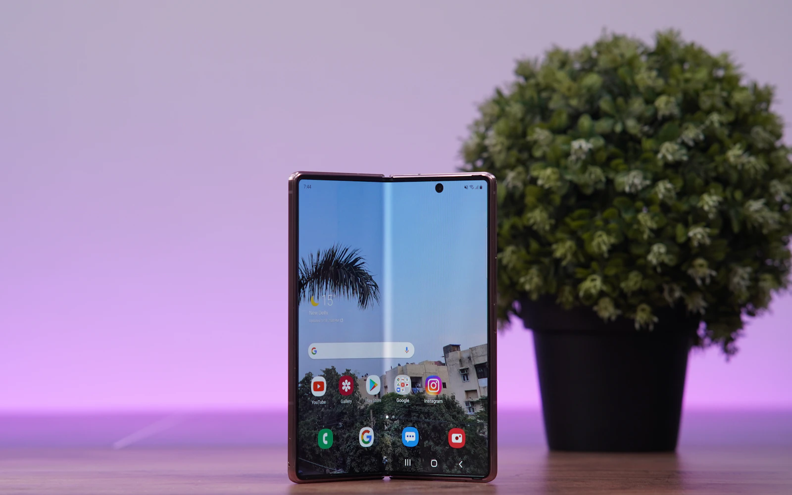 A Samsung Galaxy Z Fold standing half open on a desk beside a potted plant, its inner display showing the home screen