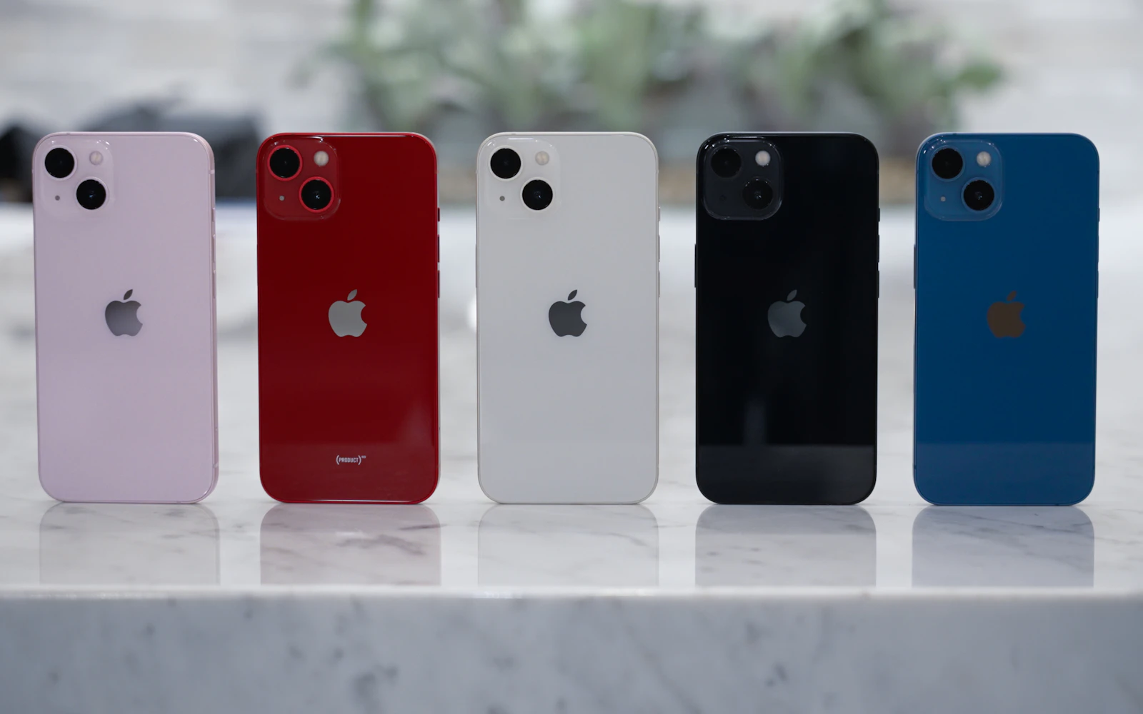 Five iPhones in pink, red, white, black and blue standing in a row on a marble counter