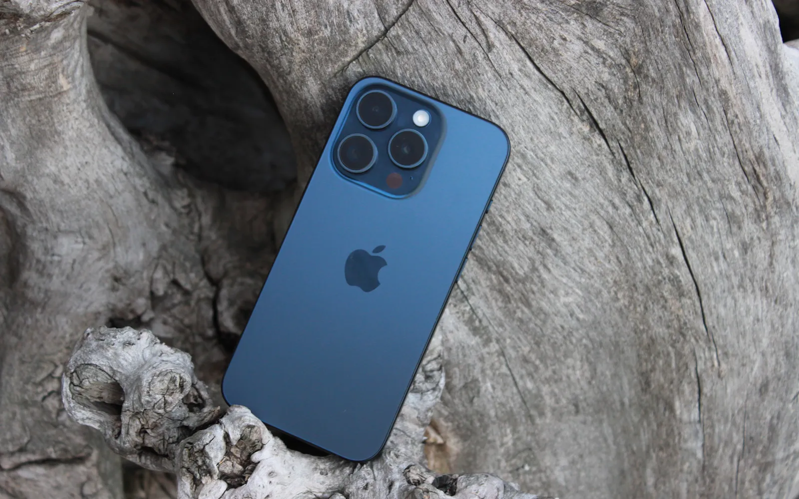 The back glass and triple camera of a blue titanium iPhone Pro lying on driftwood