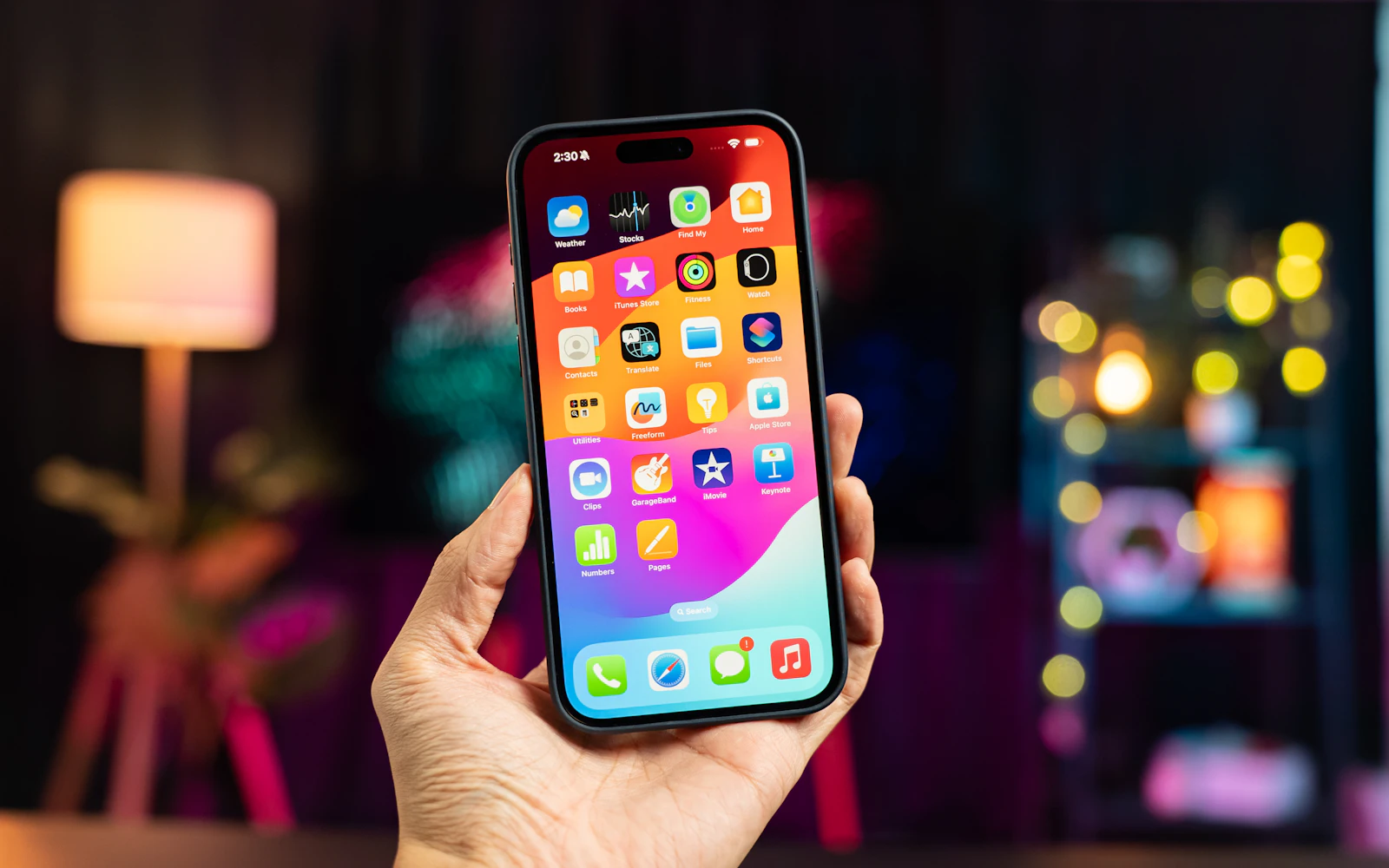 A hand holding up a recent iPhone with a Dynamic Island, its colourful home screen lit, in a dim room