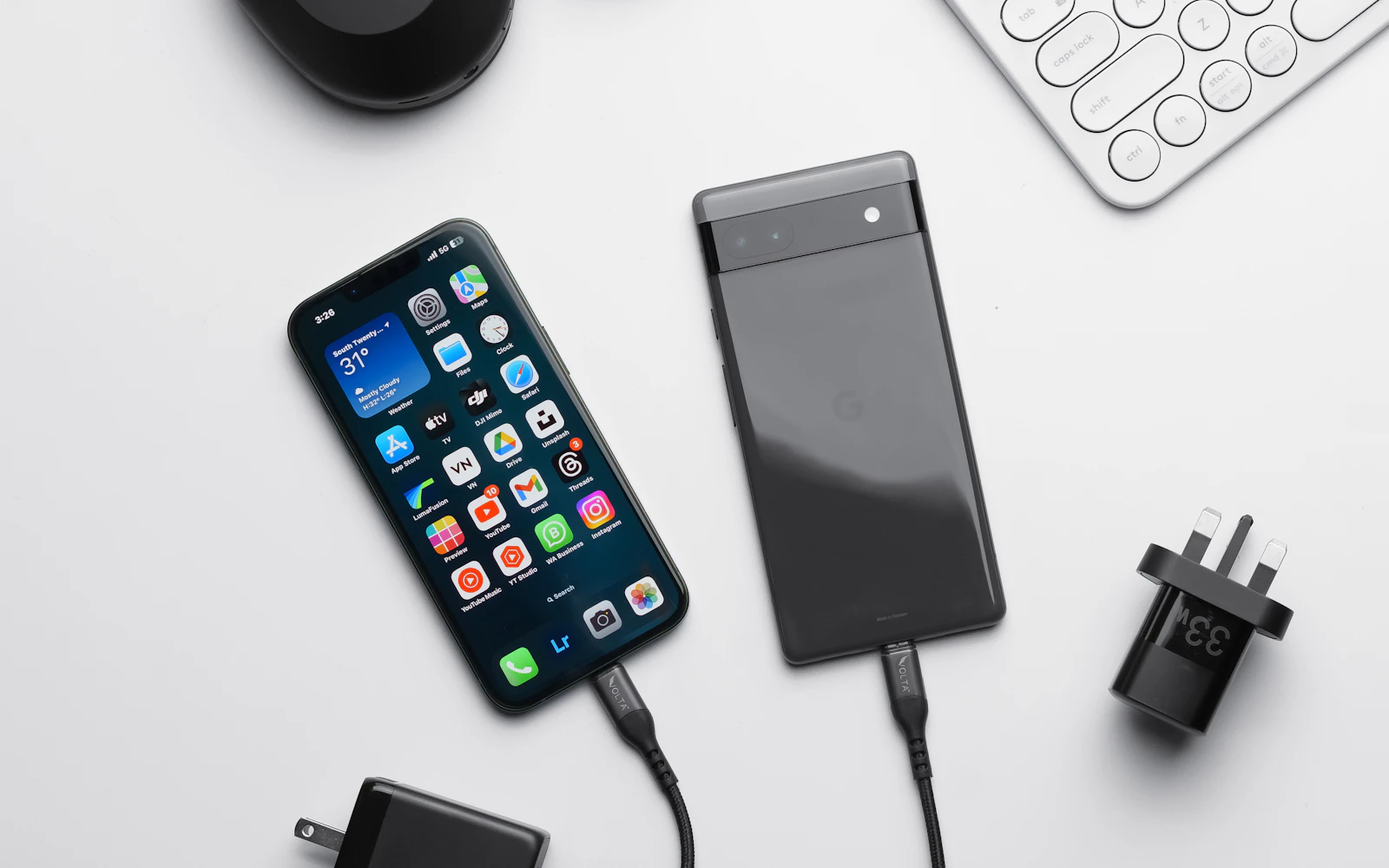 An iPhone and an Android phone on a white desk, each with a charging cable plugged into its port