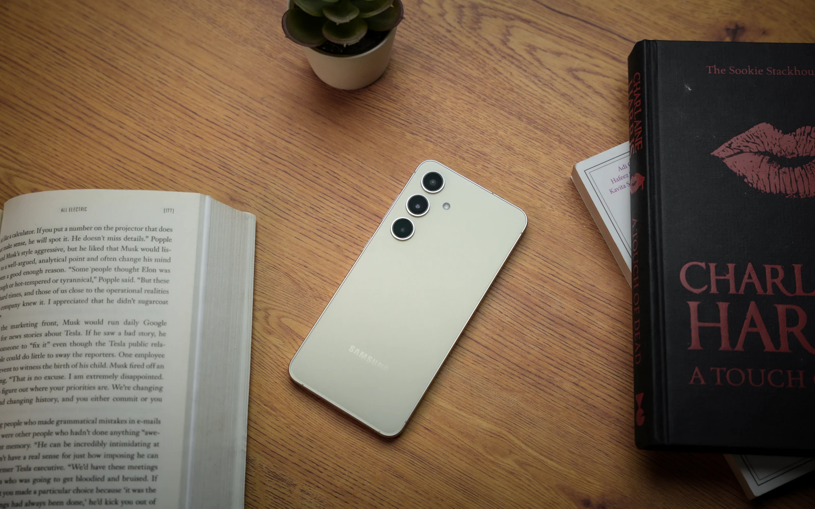 A cream Samsung Galaxy S24 lying face down on a wooden desk between books, its three rear cameras in view