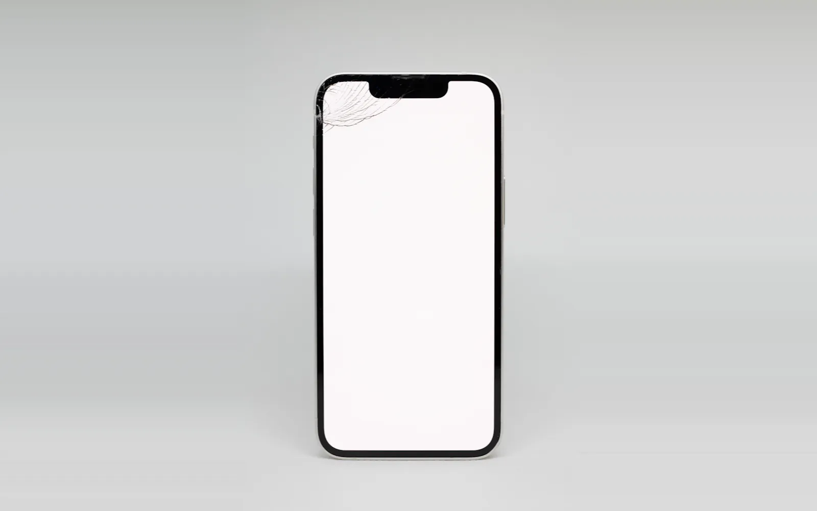 A white iPhone standing upright on a grey backdrop, its screen cracked across the top corner