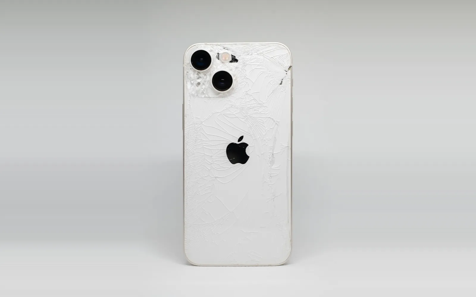 A recent iPhone with its glass back shattered across the whole panel and around the cameras, against a grey backdrop