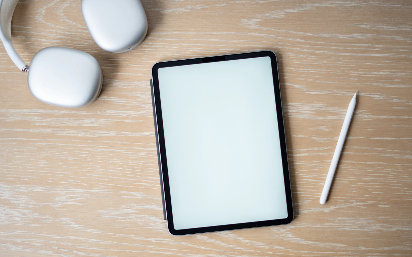 An iPad lying flat on a wooden desk beside an Apple Pencil and a pair of headphones