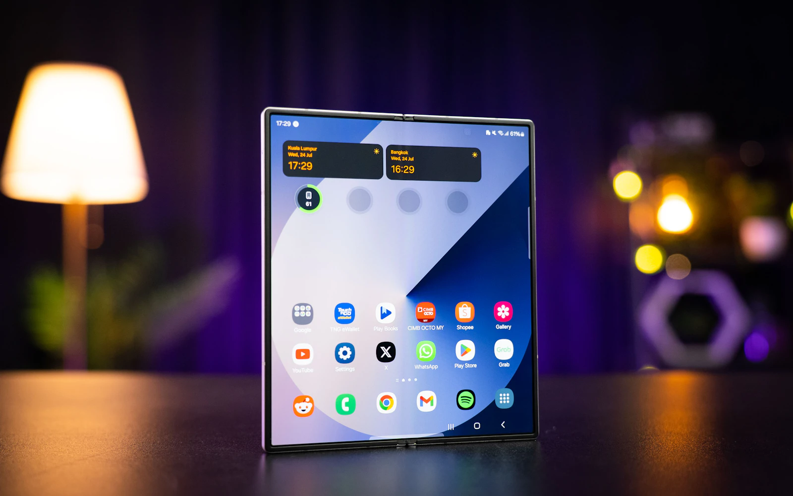 A Samsung Galaxy Z Fold standing open on a dark table, its inner screen showing the home screen, a lamp glowing behind