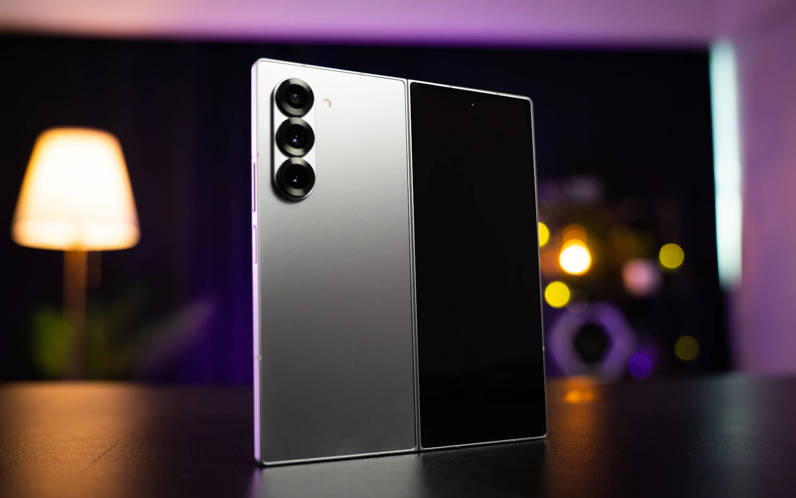 A silver Samsung Galaxy Z Fold standing upright on a dark table, its triple rear camera facing out, a lamp glowing behind