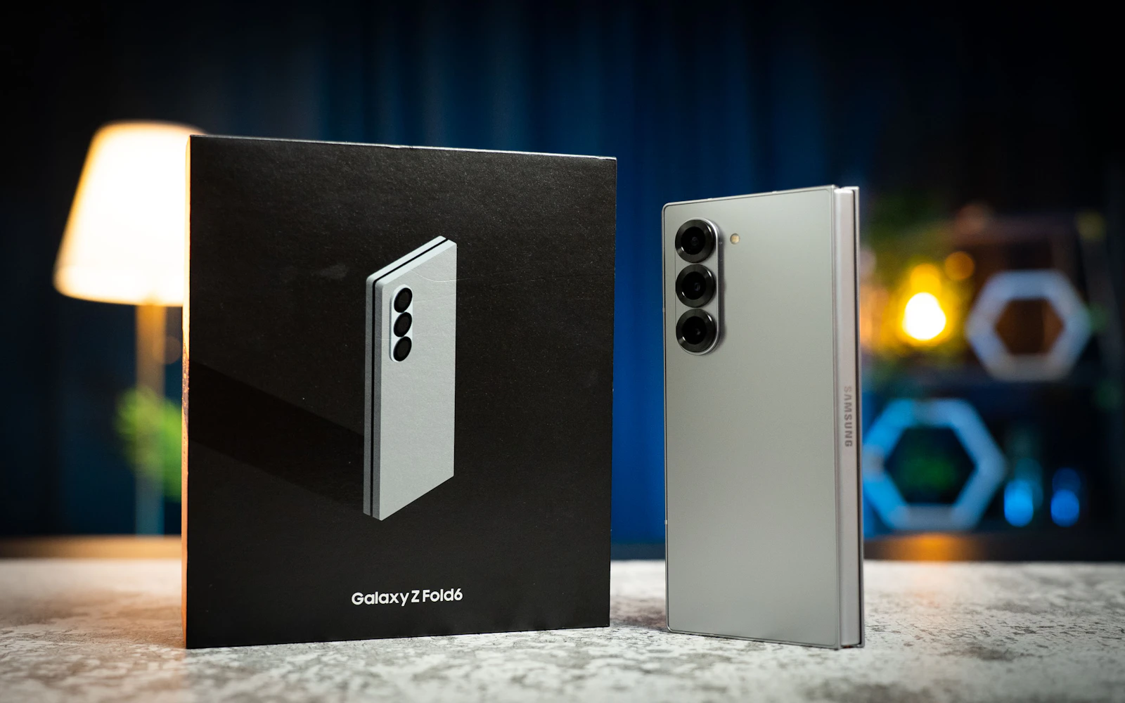 A Galaxy Z Fold6 standing folded beside its retail box on a table, a lamp glowing behind