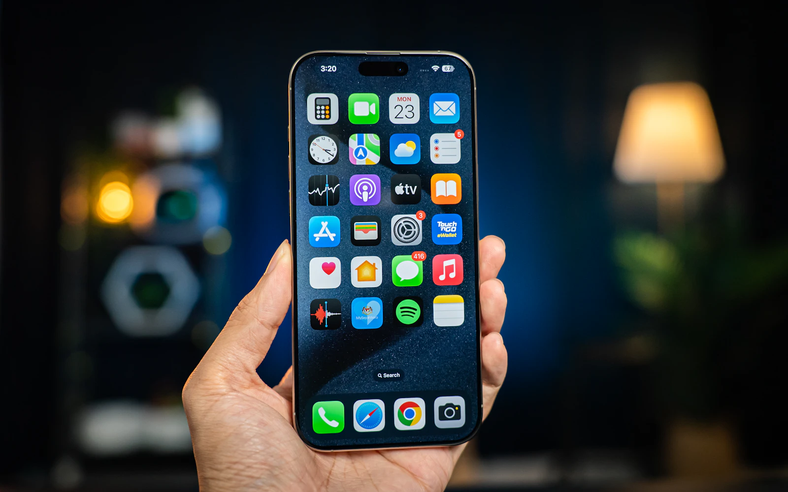 A hand holding up an iPhone Pro showing its home screen, a lamp glowing in the background