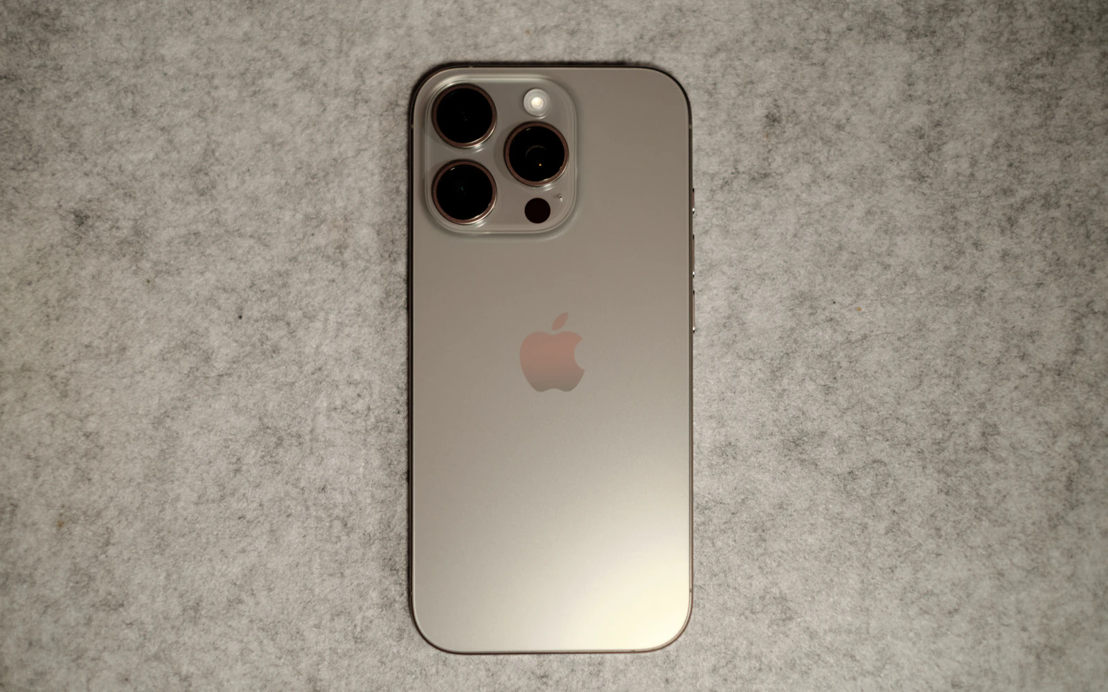 A natural titanium iPhone Pro lying face down on grey felt, its triple camera and glass back in view