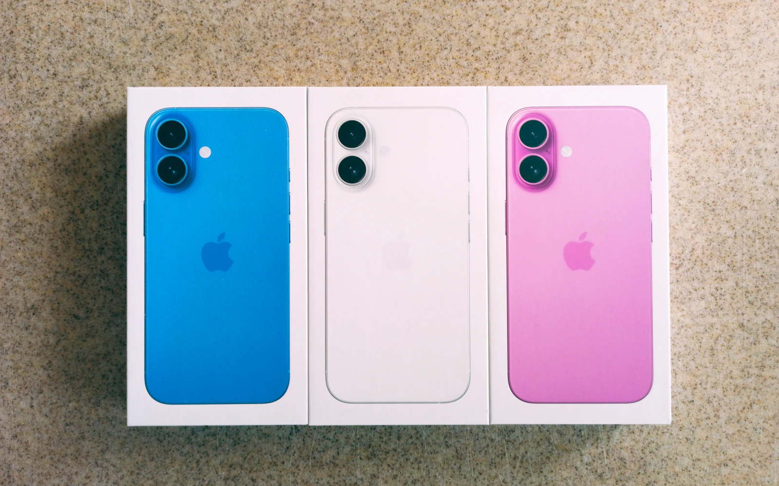 Three iPhone 16 models in blue, white and pink lying in their boxes, showing their colored glass backs