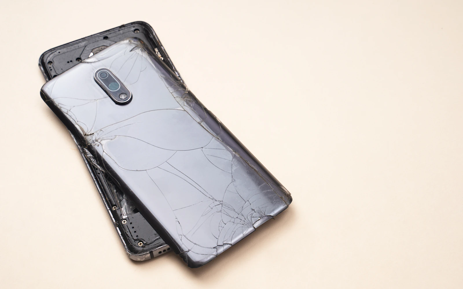 A smartphone lying on a beige surface, its glass back cracked from edge to edge