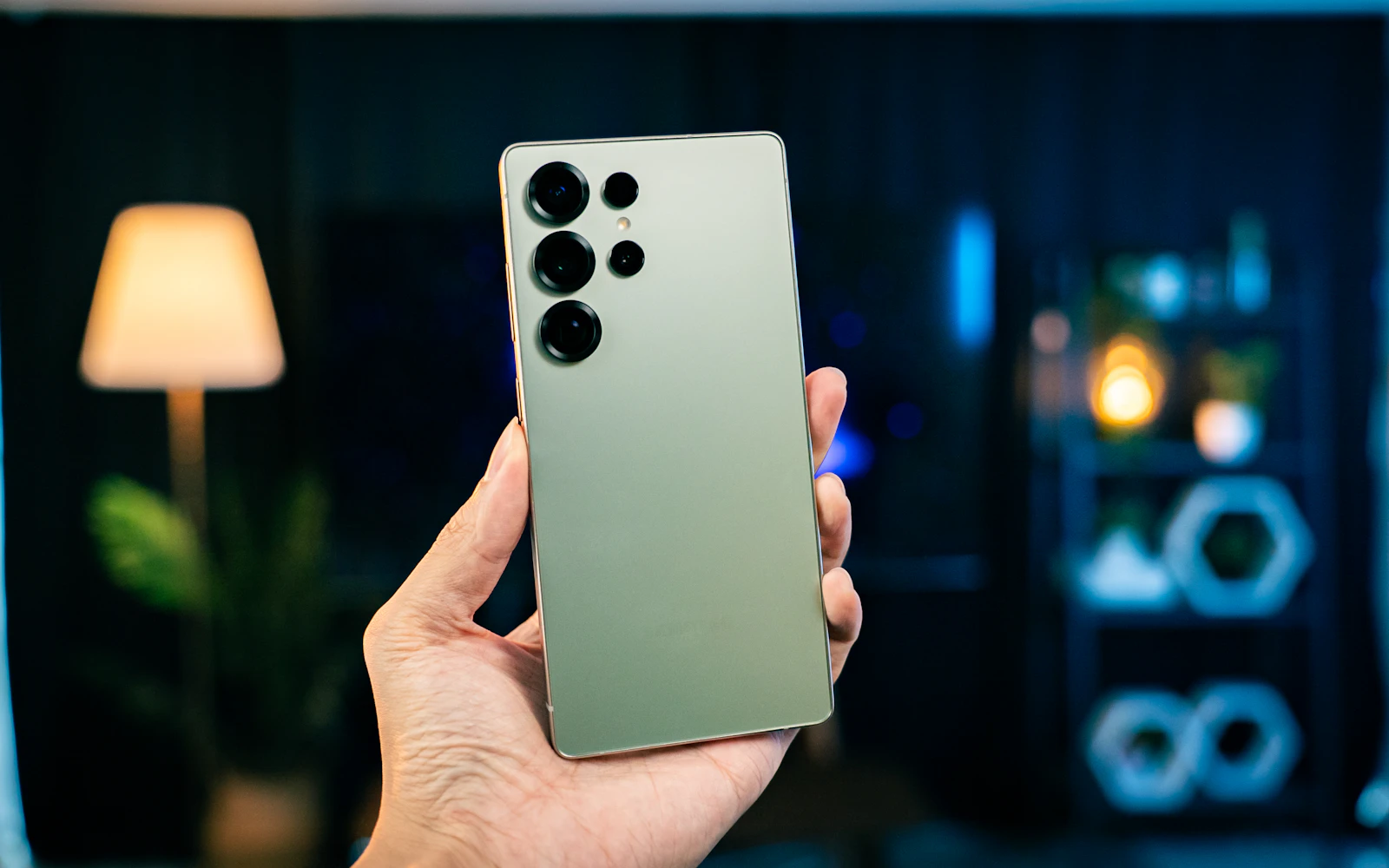 A hand holding a green Samsung Galaxy S Ultra, its rear cameras toward the lens, in a softly lit room
