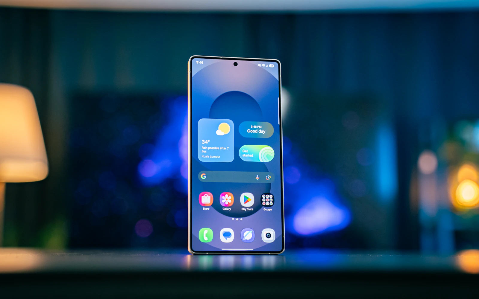 A Samsung Galaxy S25 standing upright on a table, its home screen lit, with a lamp glowing behind it