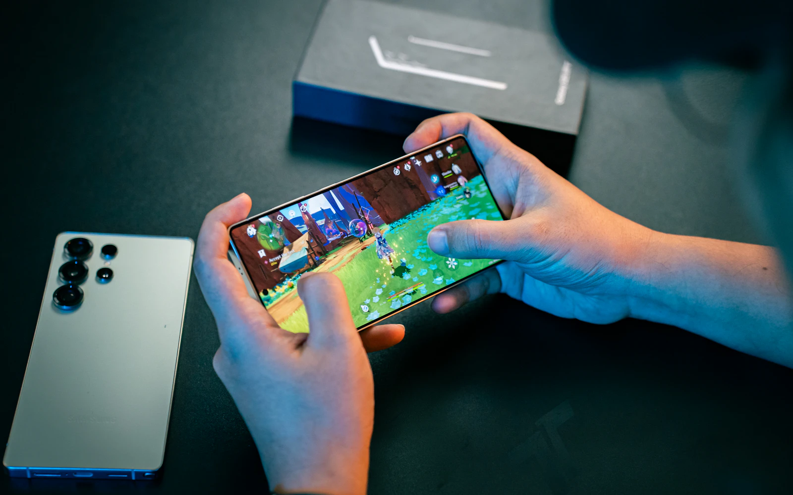Two hands playing a game on a Galaxy phone held sideways, a Galaxy S Ultra lying face down beside it
