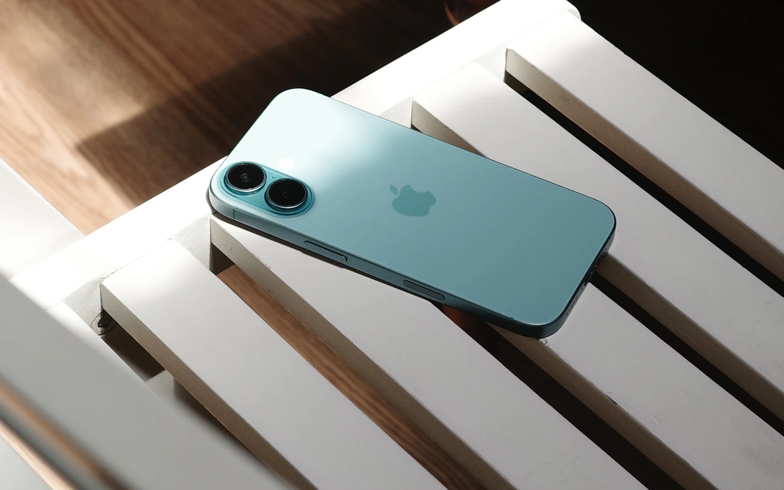 A teal iPhone lying face down on a white slatted bench in afternoon light