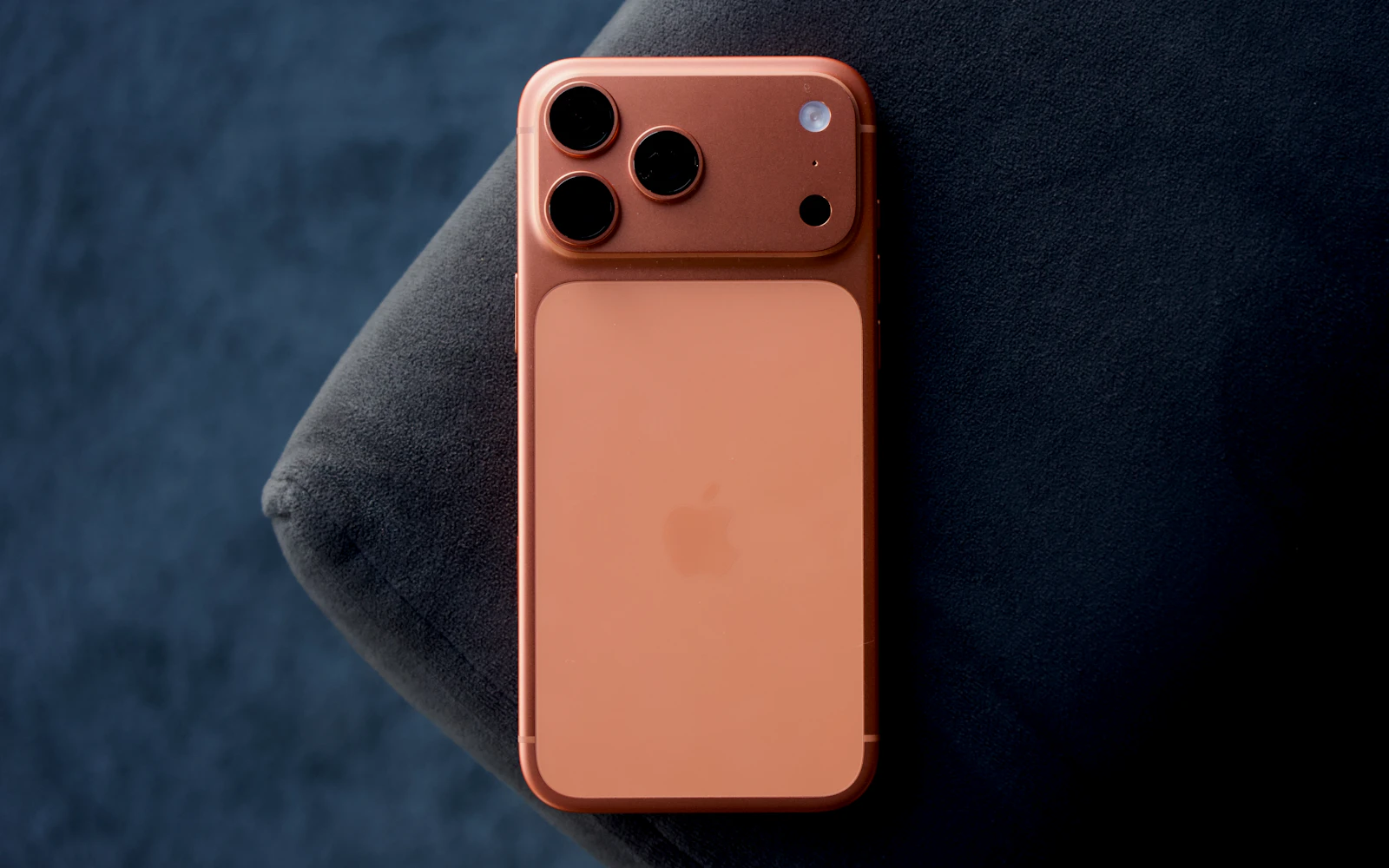 An orange iPhone Pro lying face down on a dark blue cushion, its three rear cameras in view