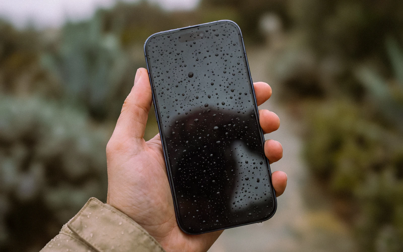 A hand holding a phone outdoors, its screen covered in water droplets