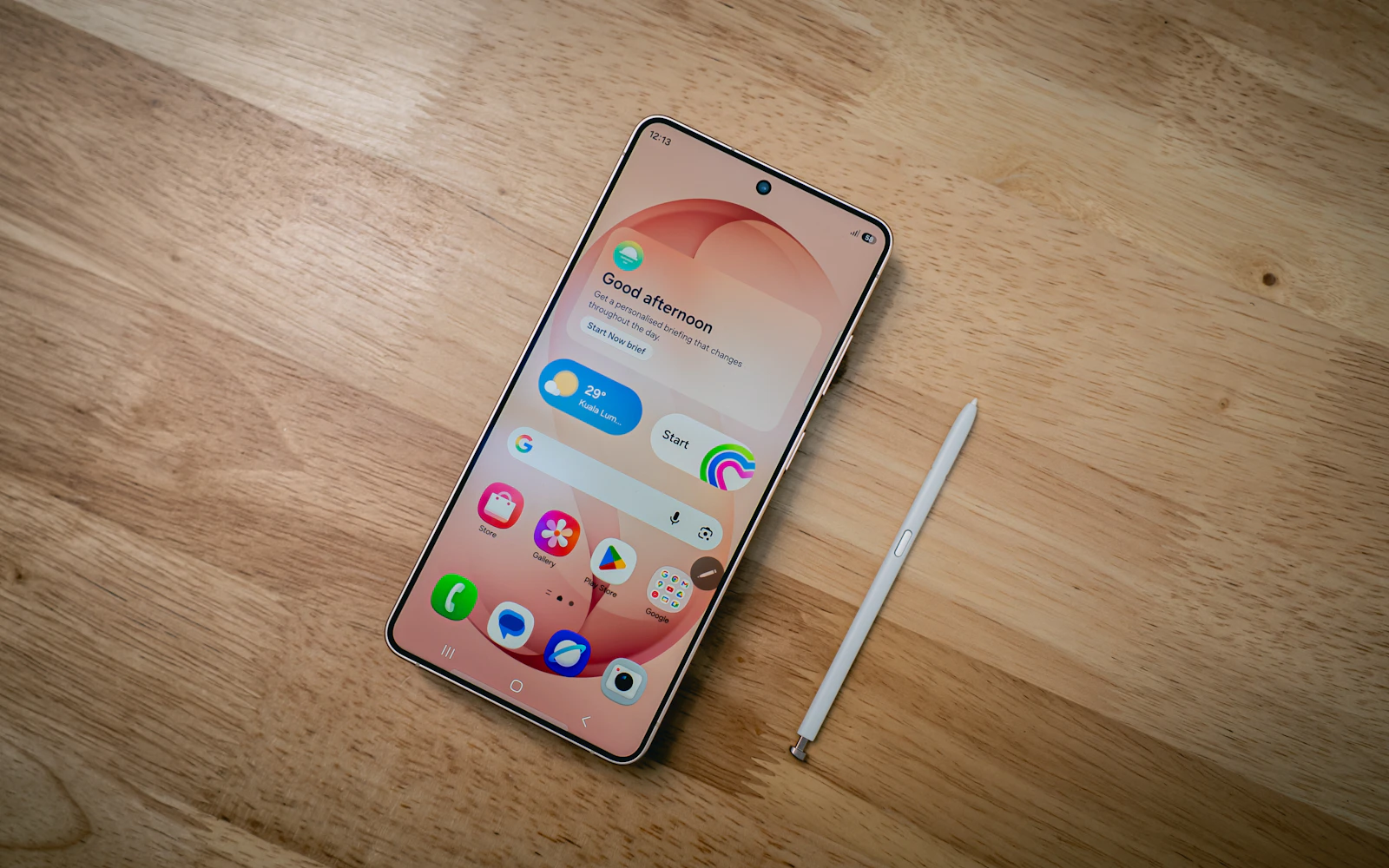 A Galaxy S Ultra phone with its home screen lit, lying on a wooden table beside its S Pen