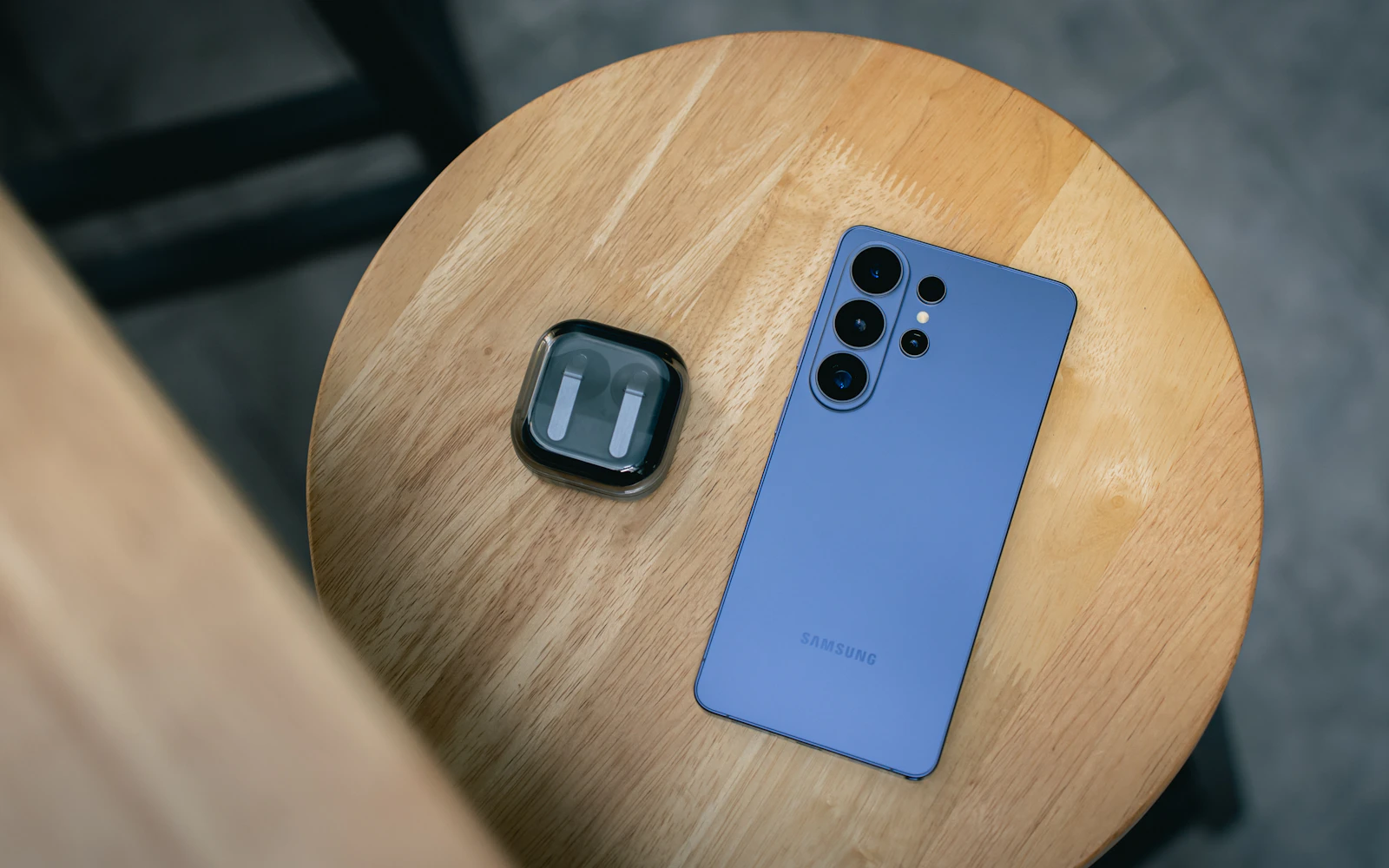 A blue Samsung Galaxy S Ultra lying face down on a round wooden stool beside a pair of earbuds
