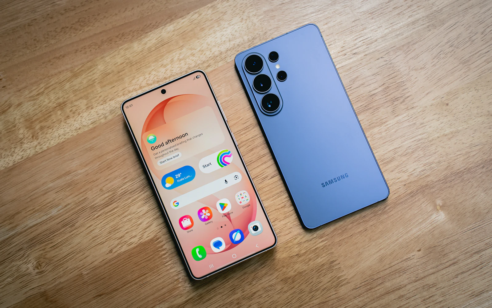 Two Samsung Galaxy phones on a wooden table, one face up showing its home screen, the other showing its blue back and rear cameras