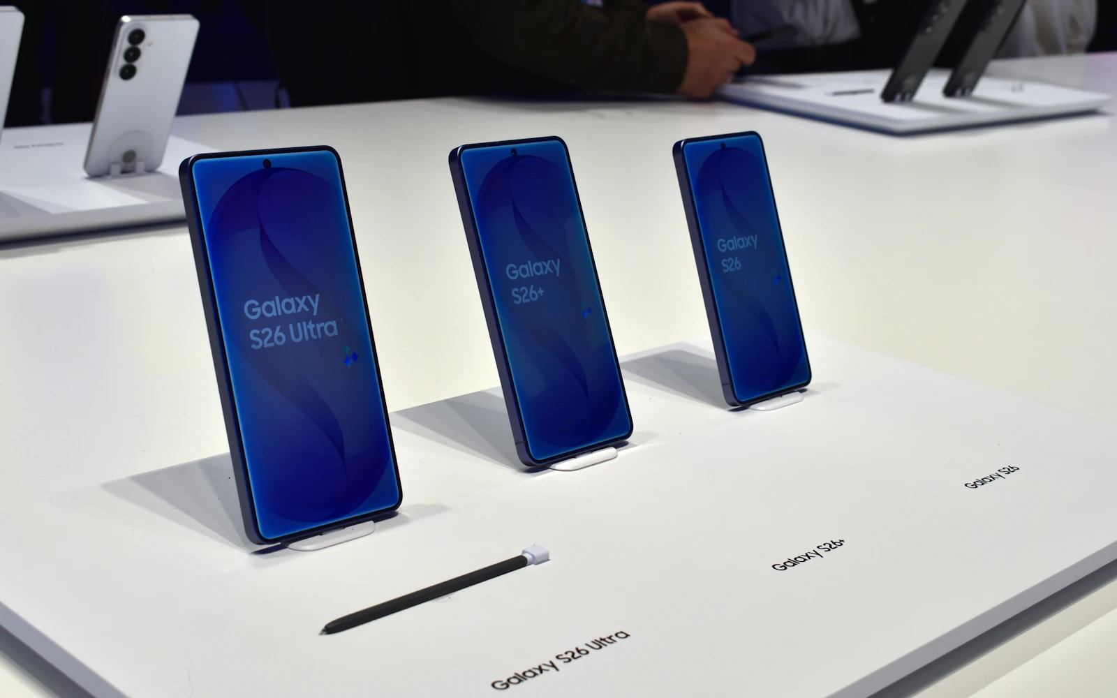 Three Samsung Galaxy S26 models, Ultra, S26+ and S26, standing on a white display table with an S Pen