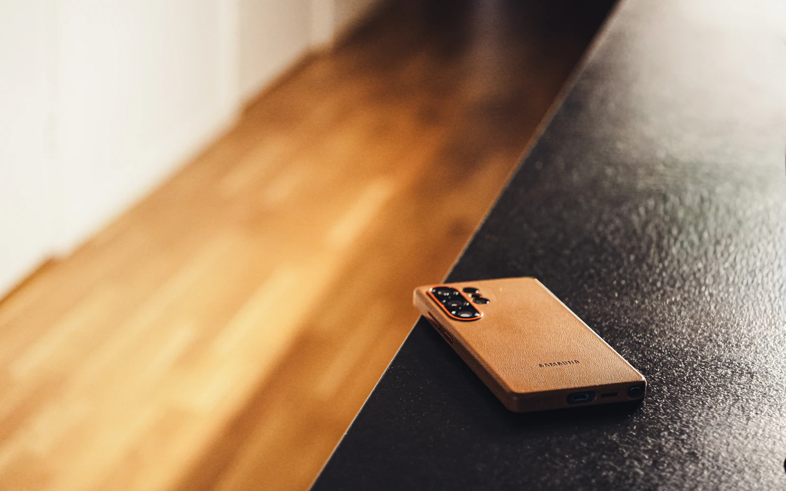 A Samsung Galaxy in a tan protective case lying face down on a dark ledge beside a wooden floor