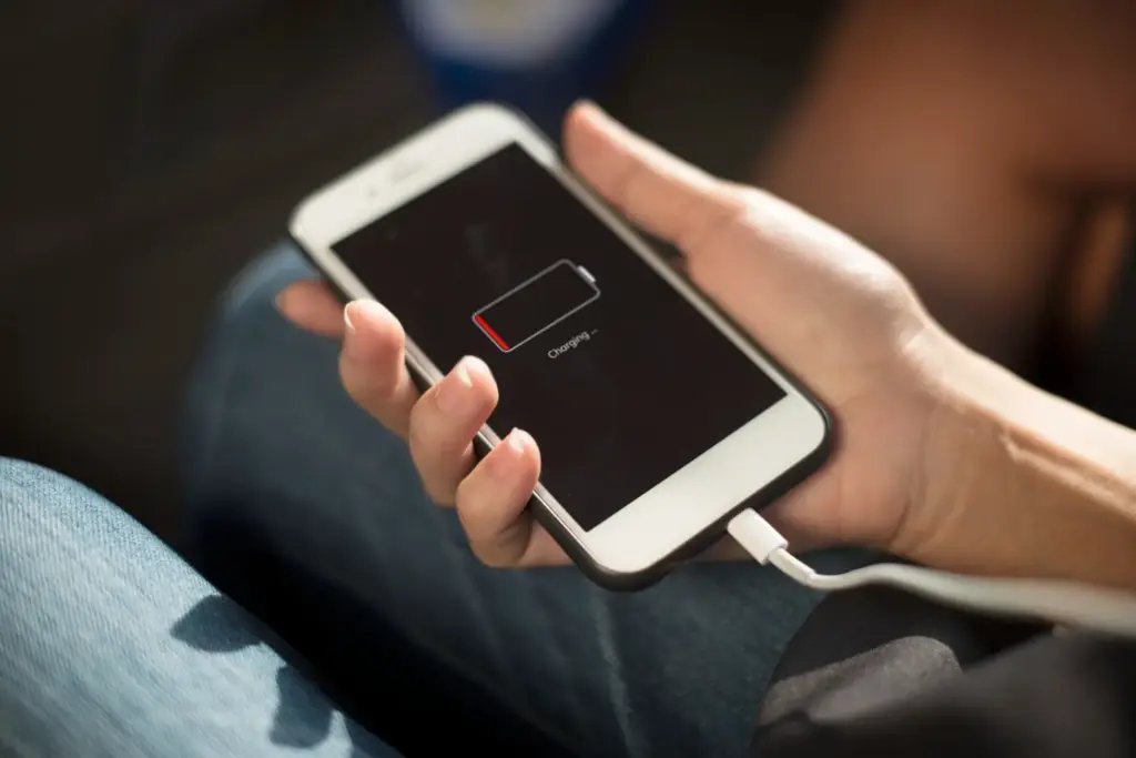 A close-up of a person holding a white smartphone connected to a lightning cable, where the screen shows a critical low battery indicator and reads 'Charging', indicating a worn-out cell that needs a professional battery replacement.